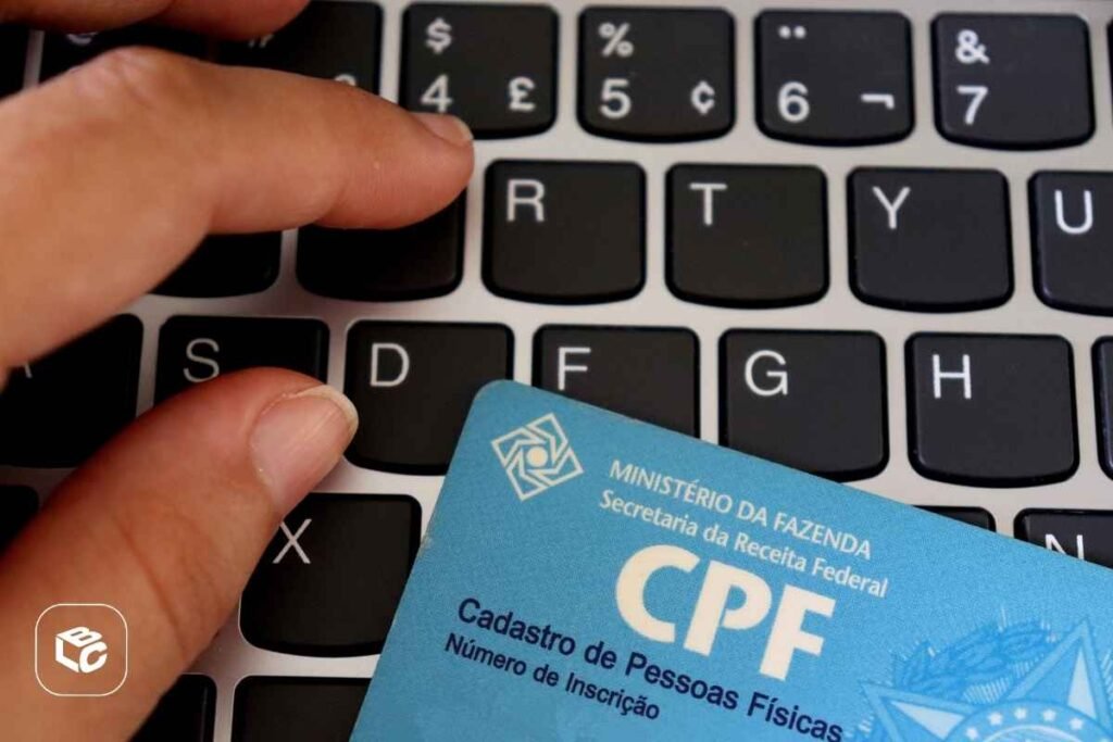 Close-up de um teclado de computador com teclas pretas e letras brancas. Sobre o teclado está parcialmente apoiado um cartão azul de CPF emitido pela Receita Federal. Uma mão aparece no canto superior esquerdo e outra na parte inferior esquerda, posicionadas próximas às teclas. No cartão é possível ler os textos “MINISTÉRIO DA FAZENDA”, “Secretaria da Receita Federal”, “CPF”, “Cadastro de Pessoas Físicas” e “Número de Inscrição”. A imagem sugere contexto de identificação cadastral, serviços digitais ou documentação fiscal. Há um pequeno logotipo branco no canto inferior esquerdo da imagem.