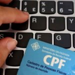 Close-up de um teclado de computador com teclas pretas e letras brancas. Sobre o teclado está parcialmente apoiado um cartão azul de CPF emitido pela Receita Federal. Uma mão aparece no canto superior esquerdo e outra na parte inferior esquerda, posicionadas próximas às teclas. No cartão é possível ler os textos “MINISTÉRIO DA FAZENDA”, “Secretaria da Receita Federal”, “CPF”, “Cadastro de Pessoas Físicas” e “Número de Inscrição”. A imagem sugere contexto de identificação cadastral, serviços digitais ou documentação fiscal. Há um pequeno logotipo branco no canto inferior esquerdo da imagem.
